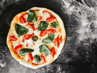 Frische belegte Pizza mit Tomaten, Mozzarella und Basilikum auf einem schwarzen Tisch mit Mehl verteilt